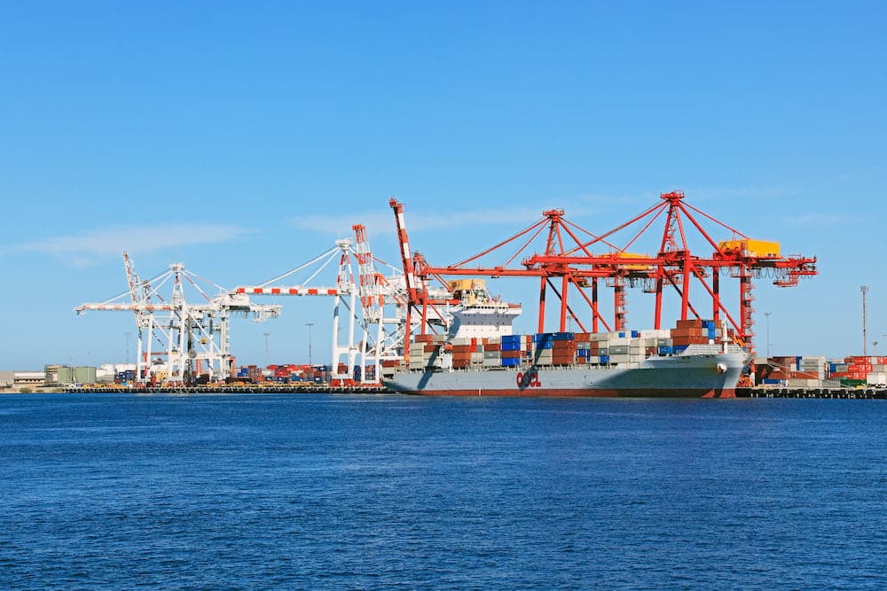 A large cargo ship is docked at a busy port in Western Australia, with cranes and containers bustling under a clear blue sky, highlighting the vibrant trade activity.