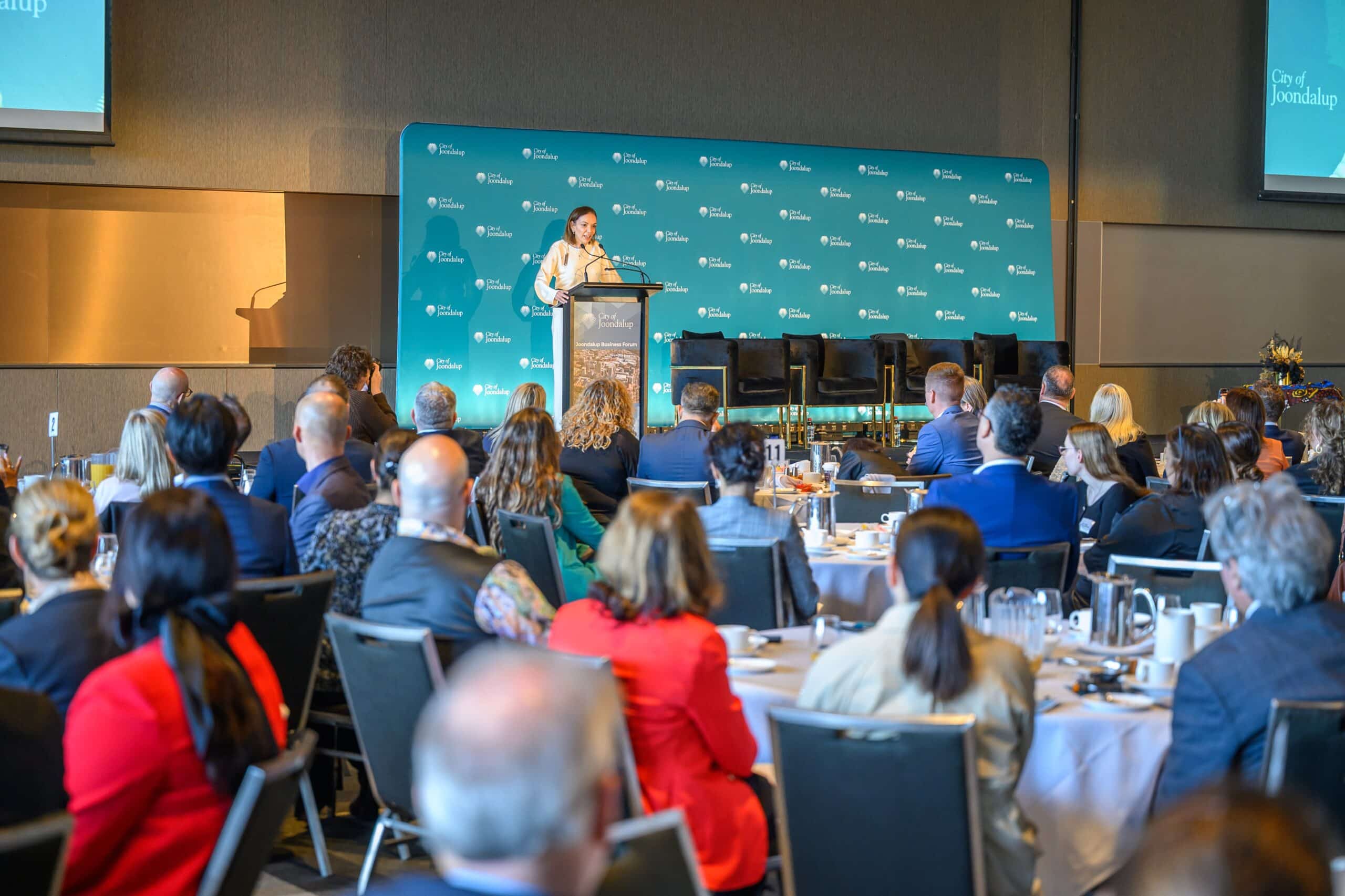 Woman standing behind a lectern delivering a presentation to people seated at tables at an event.