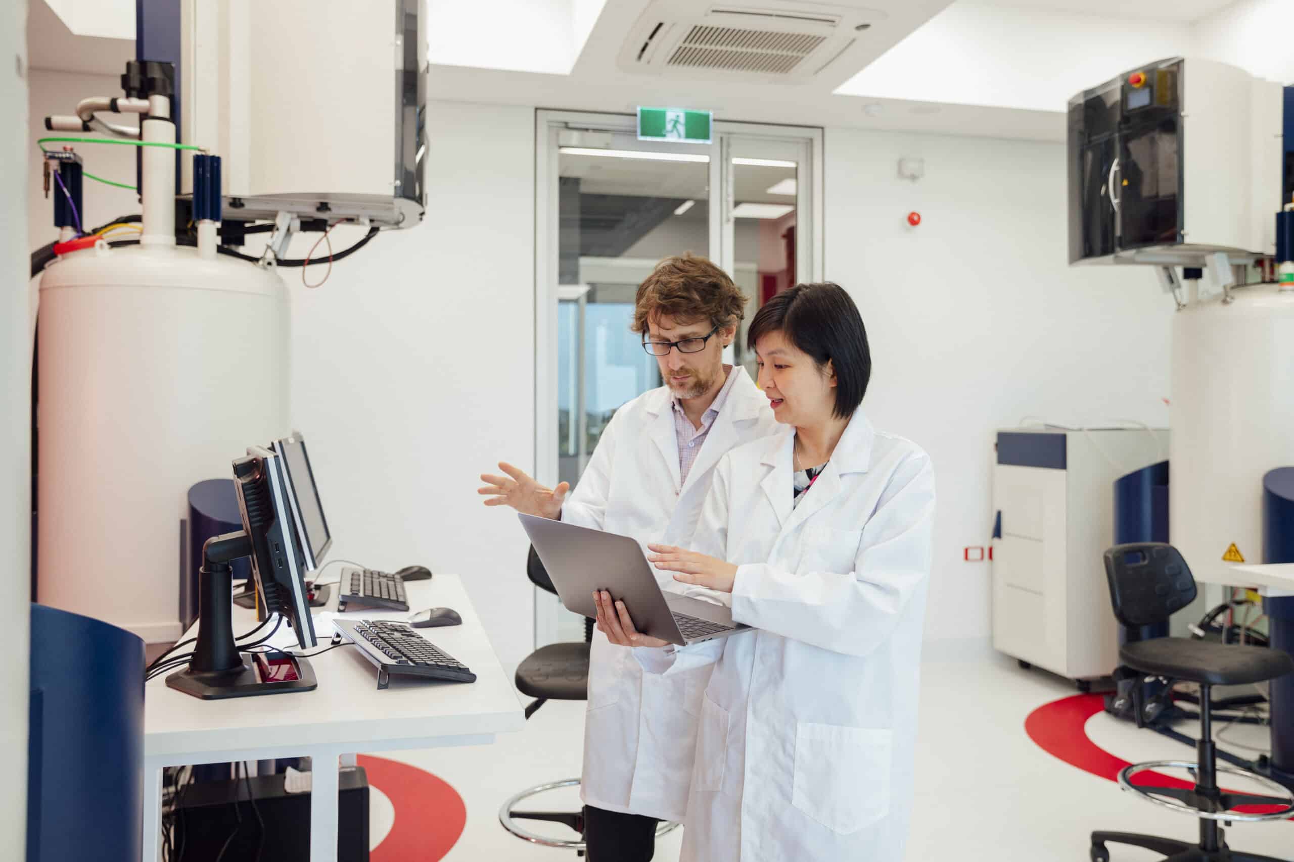Two scientists standing in a laboratory. There is a lot of large equipment surrounding them. They are wearing lab coats.